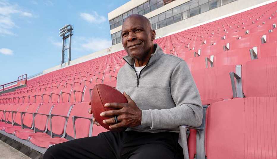 Pete Shaw, wearing a gray athletic quarter-zip, sits among red stadium seats holding a football.