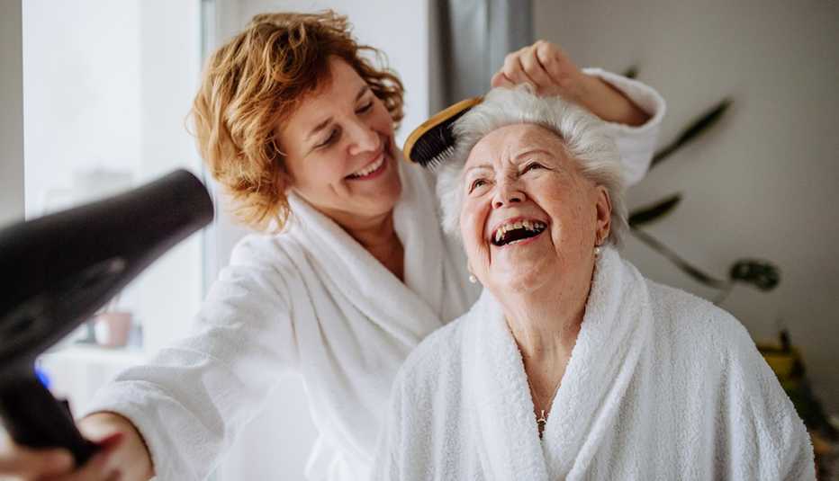two smiling women in white bath robes. one woman is blowdrying the hair of the other woman