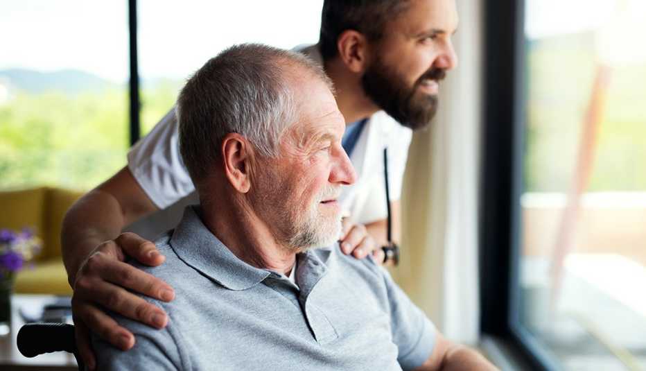 A man in a nursing home sitting in a wheel chair and looking out the window next to a male nursing homer worker