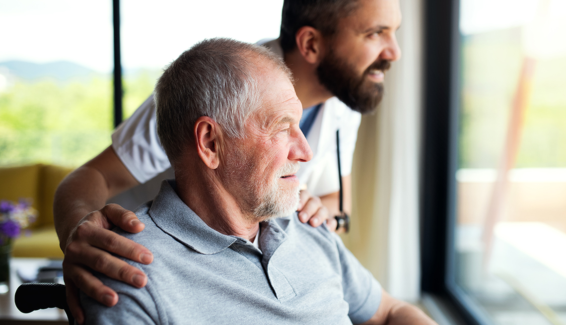 A man in a nursing home sitting in a wheel chair and looking out the window next to a male nursing homer worker