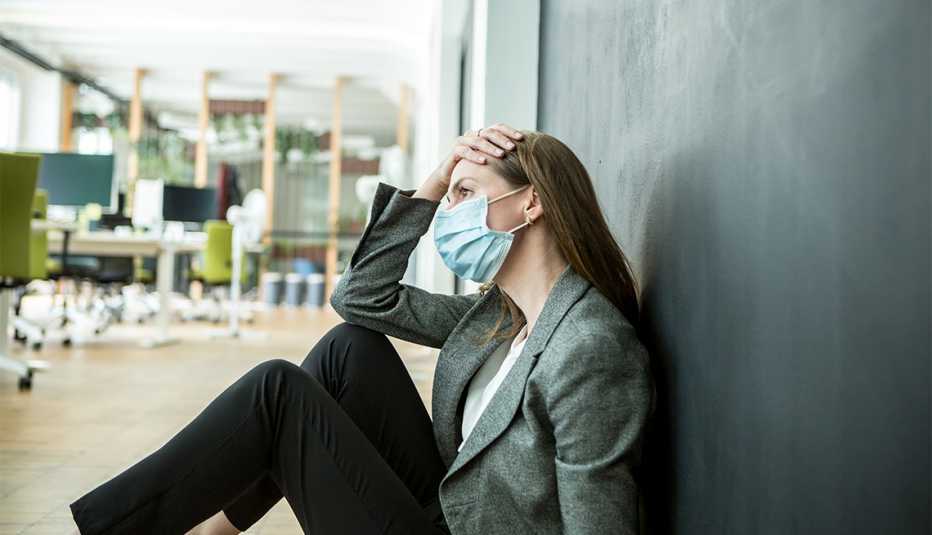 woman wearing a face mask sitting on the floor of her office with her hand on her head looking stressed