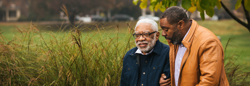 A man holding the arm of another man to help him walk in a park