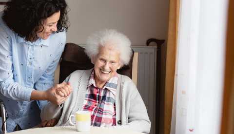 A woman is helping assist an older woman sitting in a chair in a nursing home
