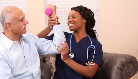 Older man does arm exercises with dumbbells with the assistance of his home healthcare physical therapist 