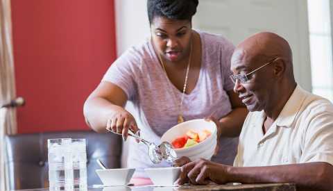 An adult daughter caregiving for her father in his home. She is serving him a bowl of fruit for lunch.
