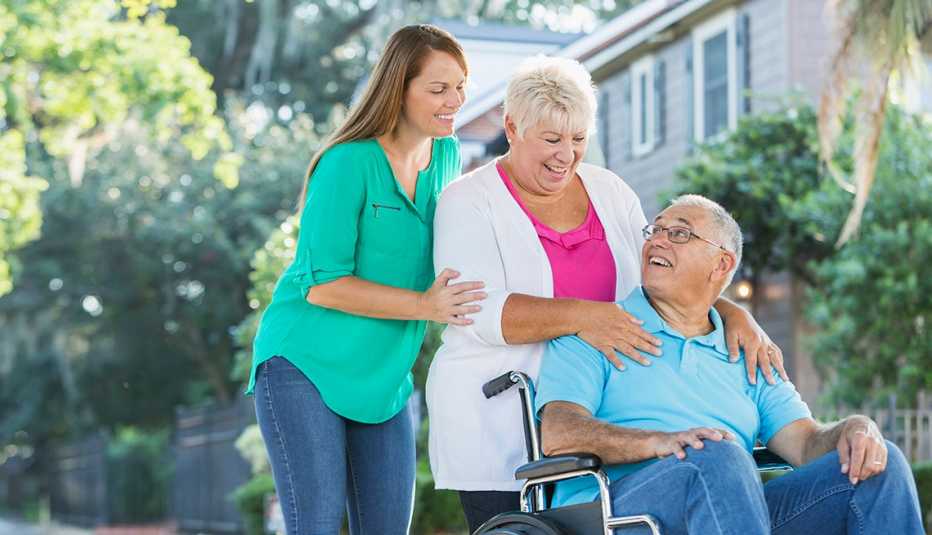 Man is sitting in a wheelchair, his wife standing behind him, hands on his shoulders. Their adult daughter is standing behind her mother
