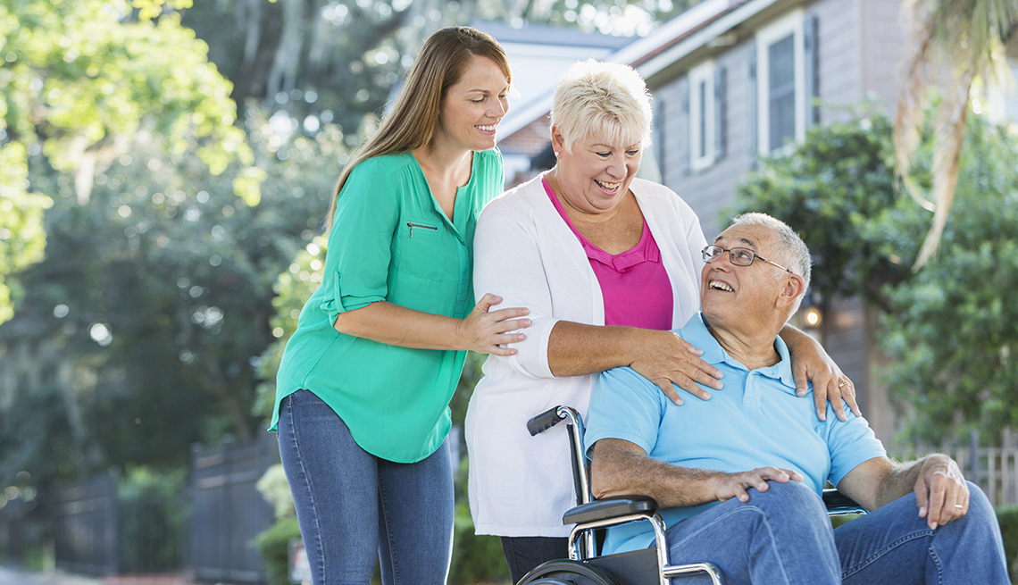 Man is sitting in a wheelchair, his wife standing behind him, hands on his shoulders. Their adult daughter is standing behind her mother
