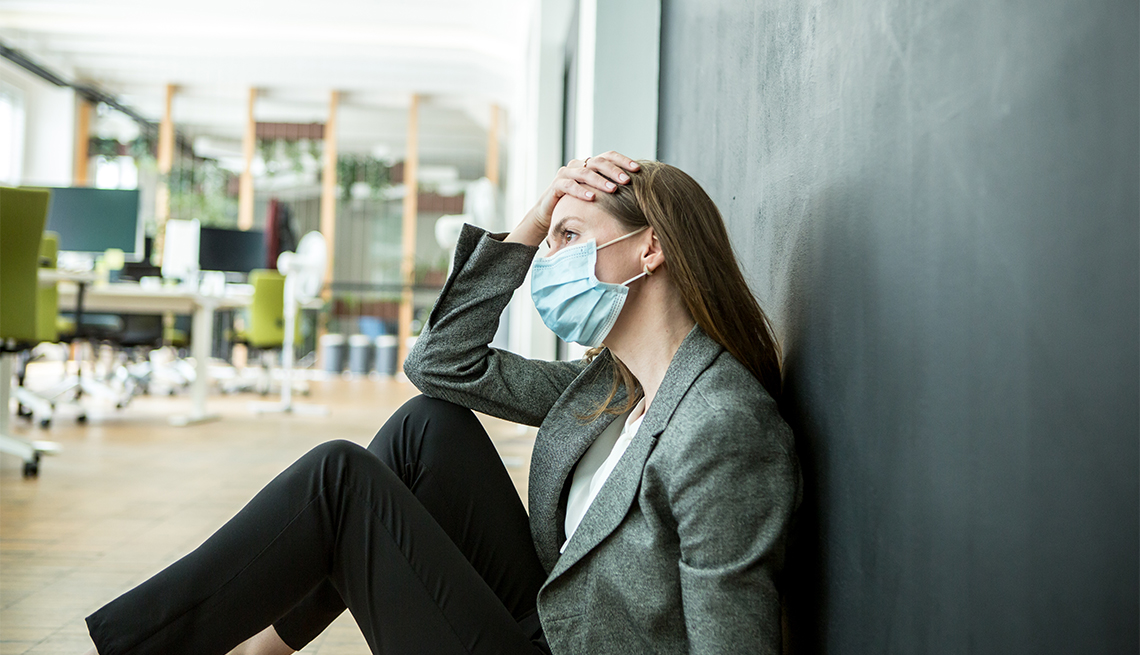 woman wearing a face mask sitting on the floor of her office with her hand on her head looking stressed