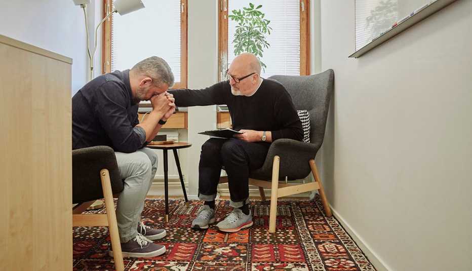 one man puts his hand on another mans shoulder to comfort him while they sit in an office setting