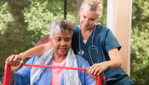 Healthcare Nurse with Patient, Long-Term Care Calculator, Caregiving, An older woman stretches an exercise band with the help of a female healthcare worker.