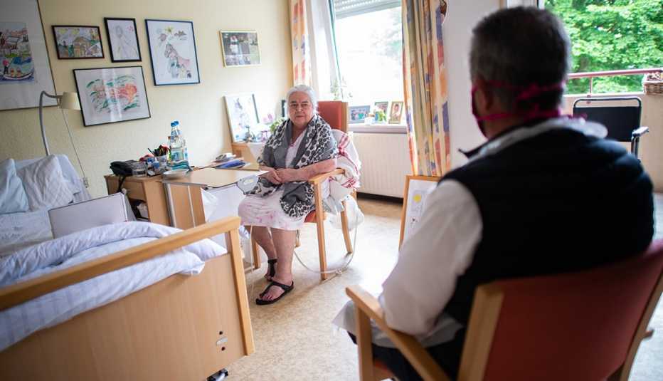 Man visiting his mother in her nursing home sitting in the chair across the room from her and wearing a face mask