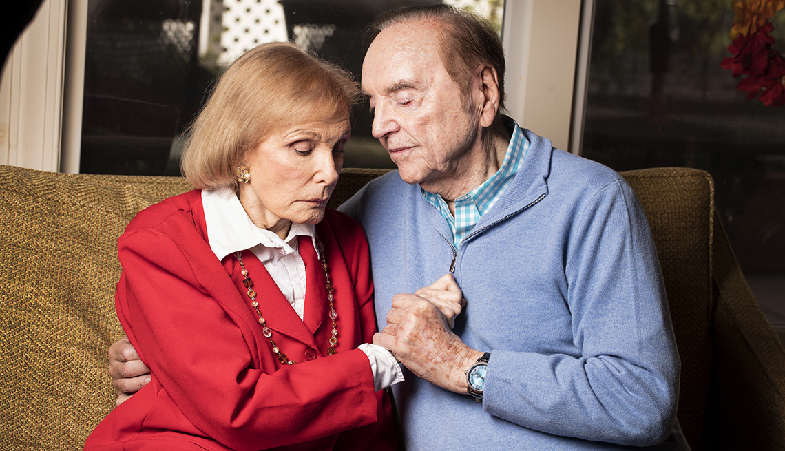 Warren Adler sits with his wife on the couch, holding hands