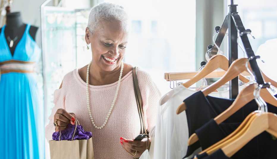 A woman shopping in a clothing store standing by a rack of dresses on hangers looking at a price tag while carrying a shopping bag