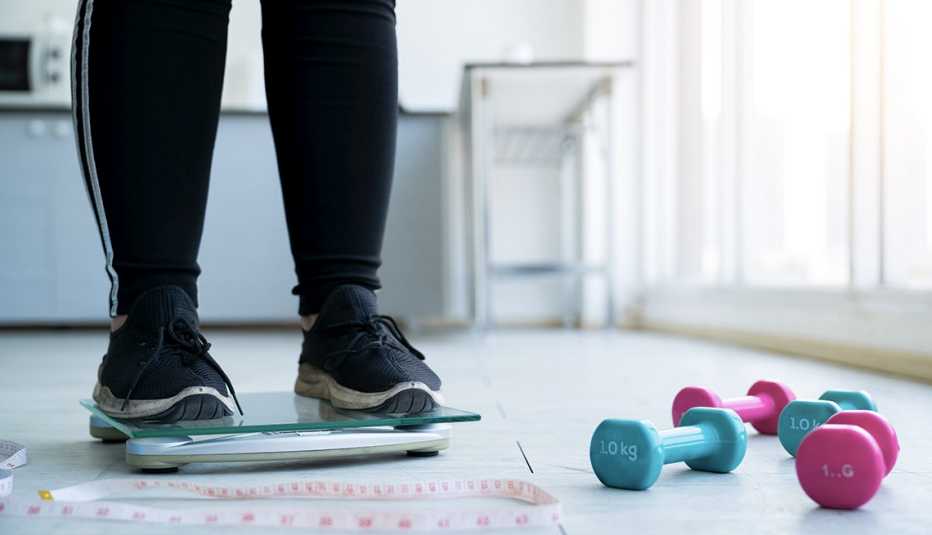 Close up legs of overweight woman checking her weight scales and drumbell, tape measure beside her.