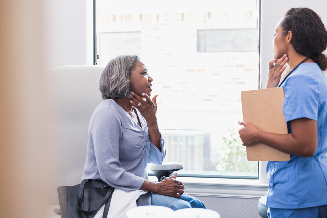 A woman gestures towards her face while talking with a dermatologist