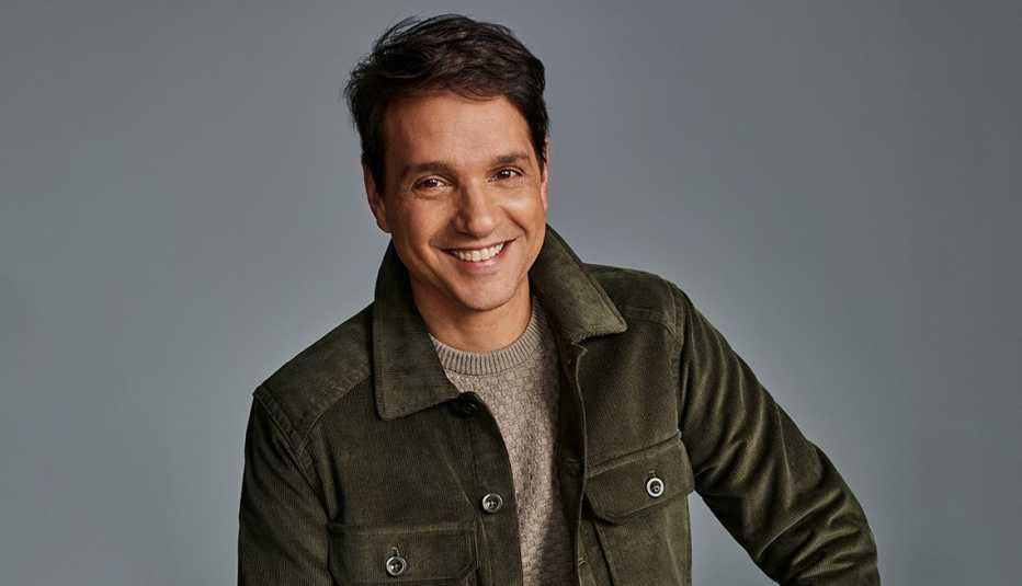 ralph macchio smiling, wearing tan shirt and brownish green jacket in front of gray background