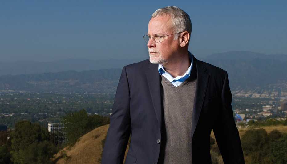 michael connelly with head turned over his right shoulder on top of mountain with buildings and trees in the distance behind him