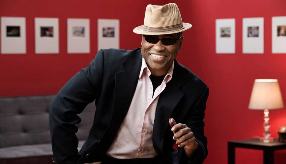 A man smiling while wearing a hat sunglasses suit jacket and collared shirt