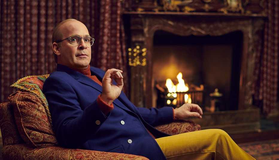 Tom Hollander sitting in a chair nearby a fireplace as his character Truman Capote in "Feud: Capote Vs. The Swans."