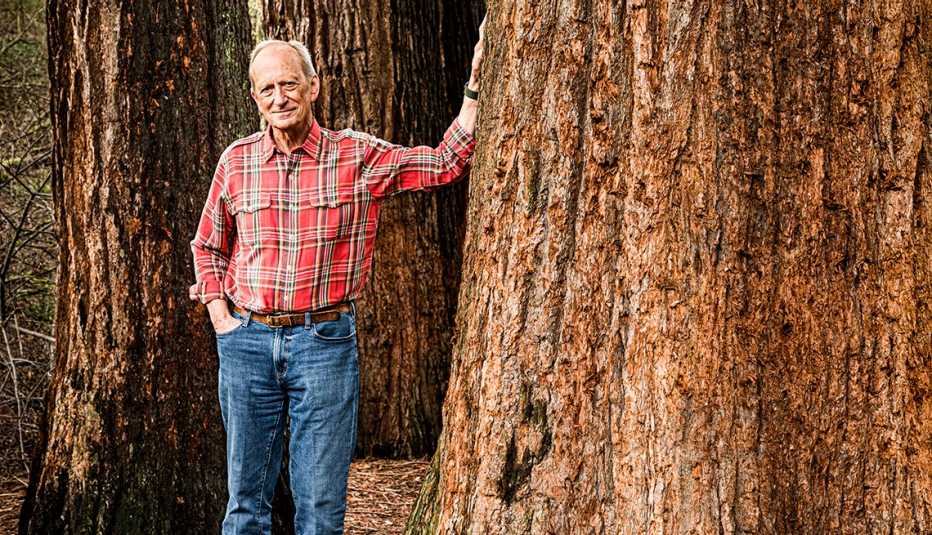 denis hayes poses in a forest with his hand on a large tree trunk
