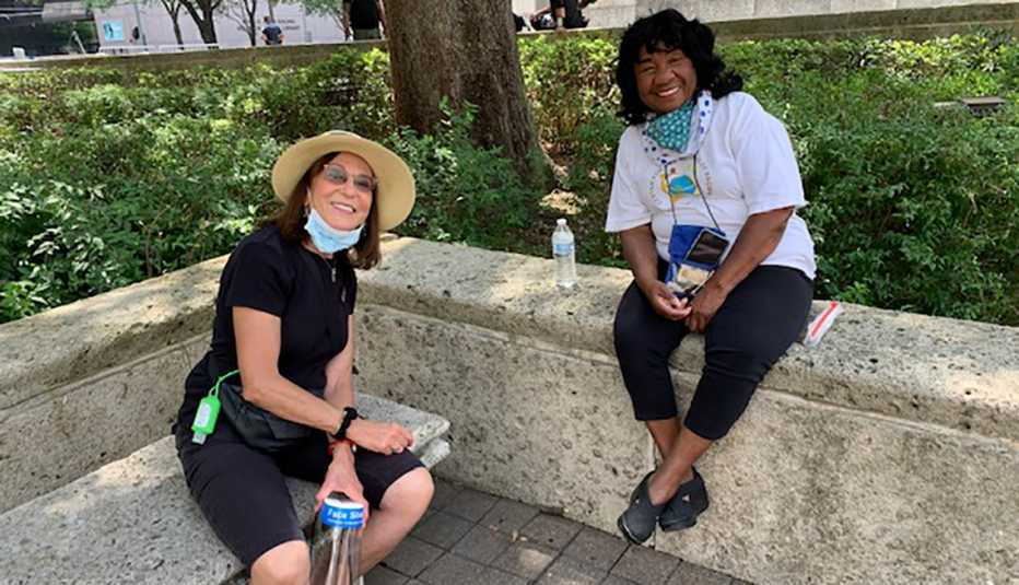 two women have stopped to rest on a city bench and low marble wall and are smiling at the camera with their covid face masks down two women have stopped to rest on a city bench and low marble wall and are smiling at the camera with their covid face masks down