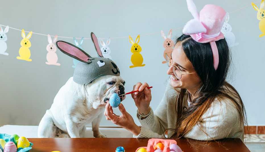 woman paints easter eggs while a bulldog in a bunny hat watches