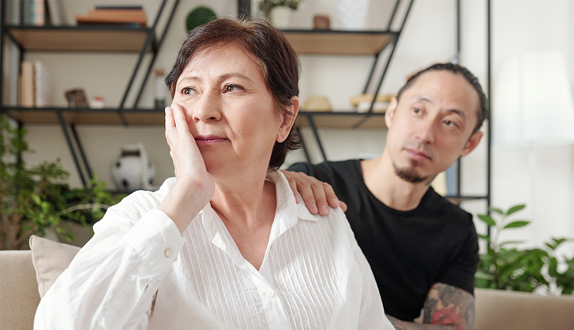 Sad mature mother sitting on sofa and looking away with her son sitting in the background and supporting her