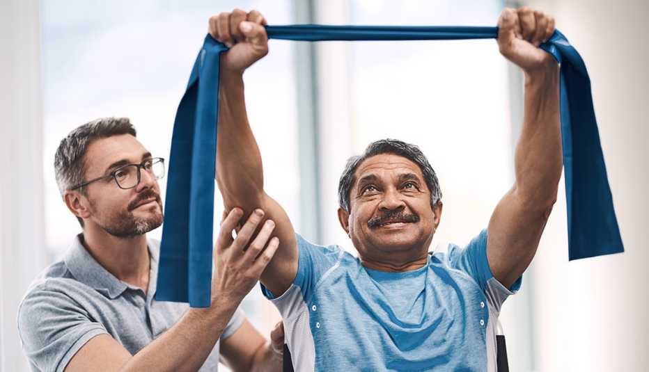 a man stretches blue exercise bands overhead for joint pain relief with the help of a physical therapist