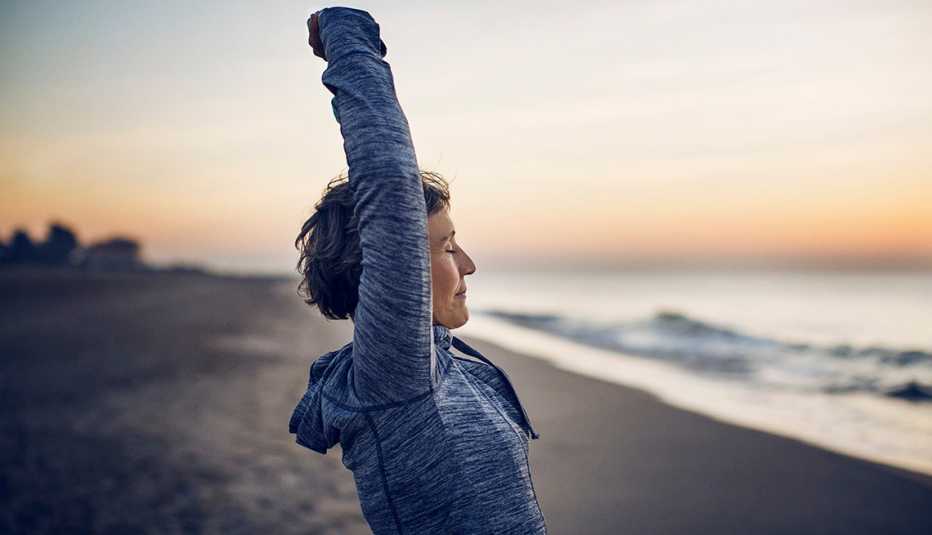 older woman stretching overhead on a beach at dusk