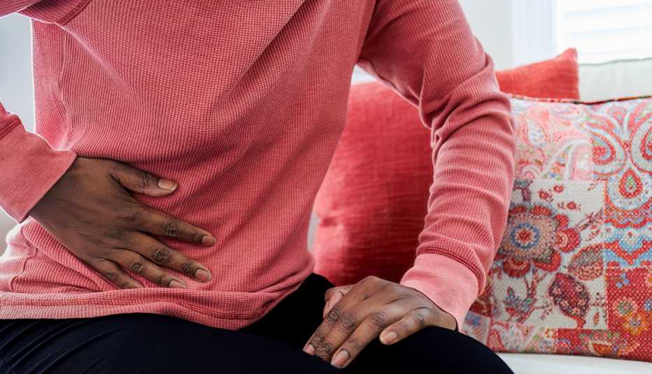 woman in a pink sweater holding her side in pain from stomach flu or food poisoning while sitting on a couch with pink patterned pillows