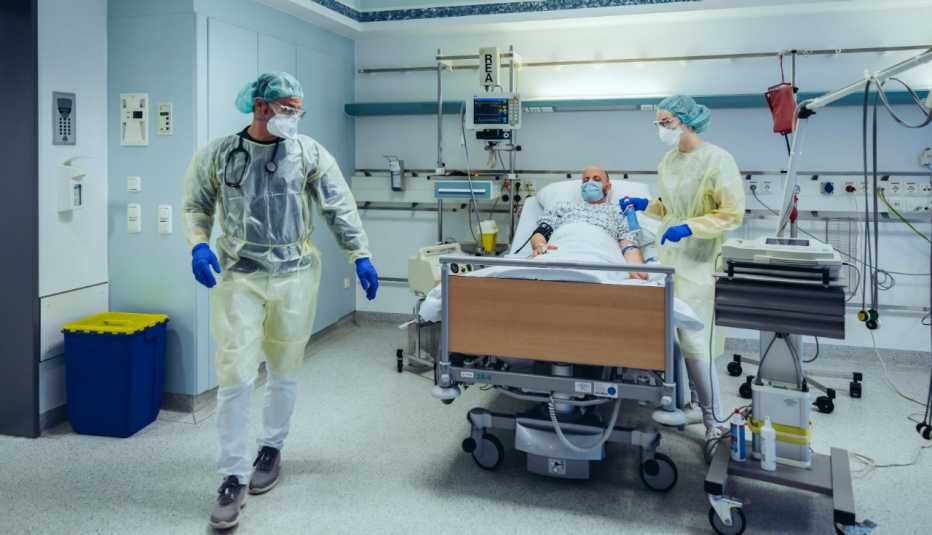 hospital workers in masks and protective wear attending to patient lying in bed, wearing mask