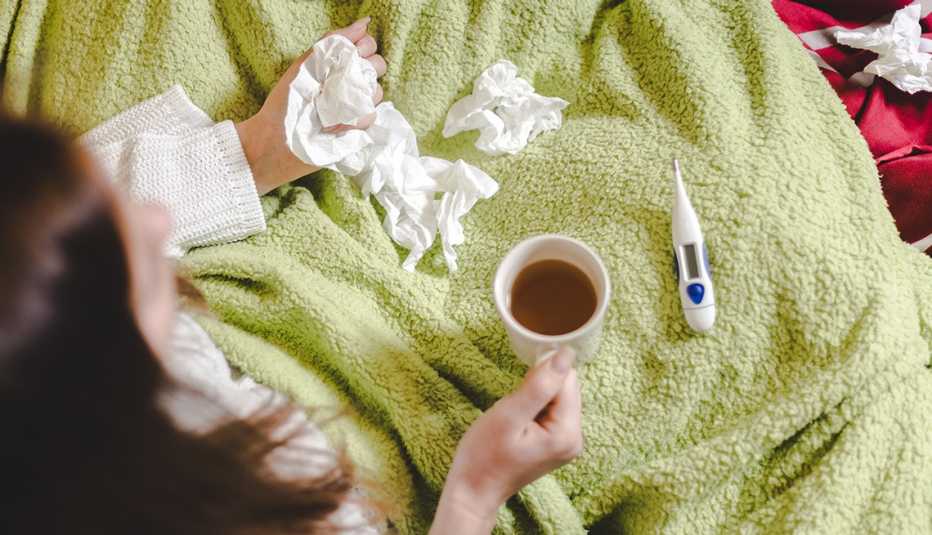 overhead photo of woman sitting up in bed holding a hot cup of tea with a thermometer and used tissues on her lap