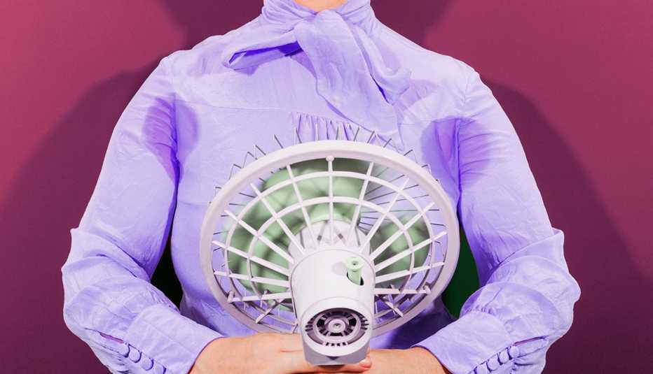 closeup of womans torso she is holding up a small fan to her body and wearing a long sleeve blouse that has sweat marks under the armpits