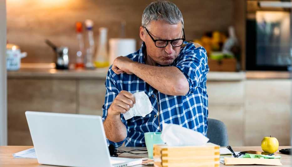 Man sneezing into his elbow. Laptop and tissues are in front of him.