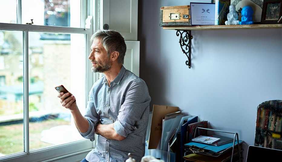 Man looking out window as he holds a smart phone. 