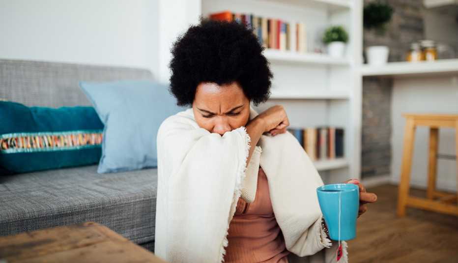 woman home wrapped in a blanket coughing and holding a cup of tea