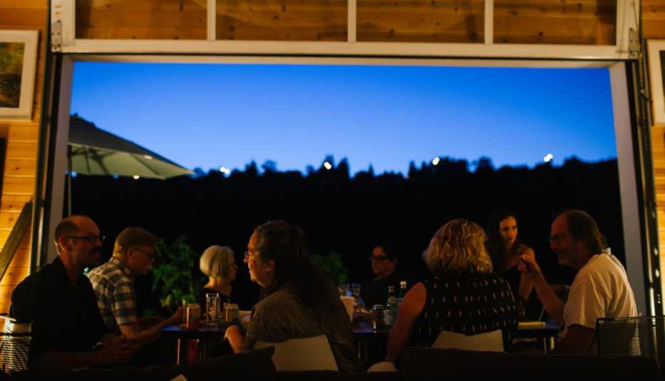 A group of eight people are seated at a table enjoying a meal next to an open garage door that shows a view of an evening sky