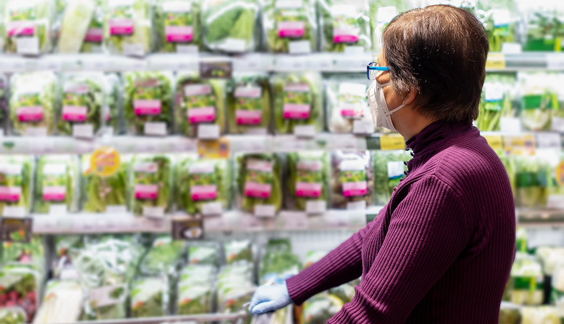 a woman wearing a face mask and gloves pushes a cart in a grocery store in an aisle of bagged salad