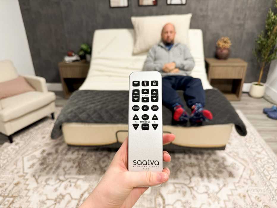The remote control of the Saatva Adjustable Base held up in front of the bed where a man is sitting.