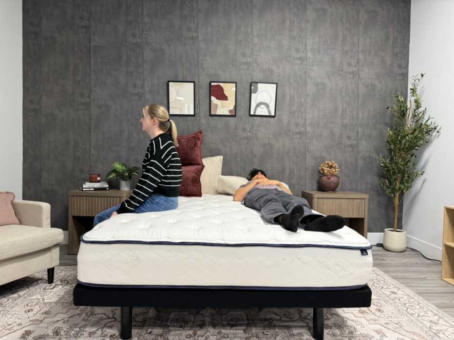 A woman sitting on the edge of a mattress while another person lies on the opposite side