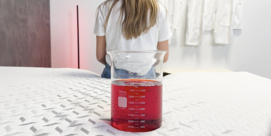 Woman in background sitting on edge of mattress while in the foreground sits a beaker filled with red liquid balancing on the bed

