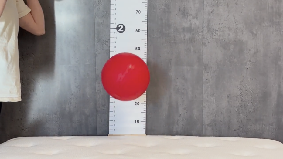 A red ball bounces on a white mattress with a ruler in the background to measure how high it goes