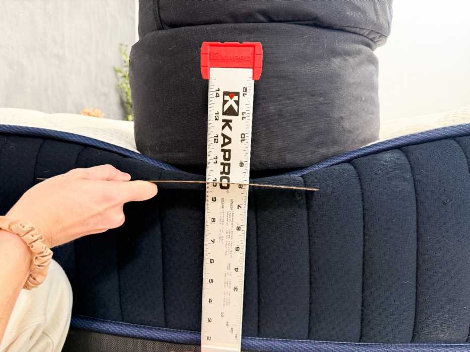 A sandbag sits on the edge of a mattress while a person measures how low the mattress sinks with a ruler
