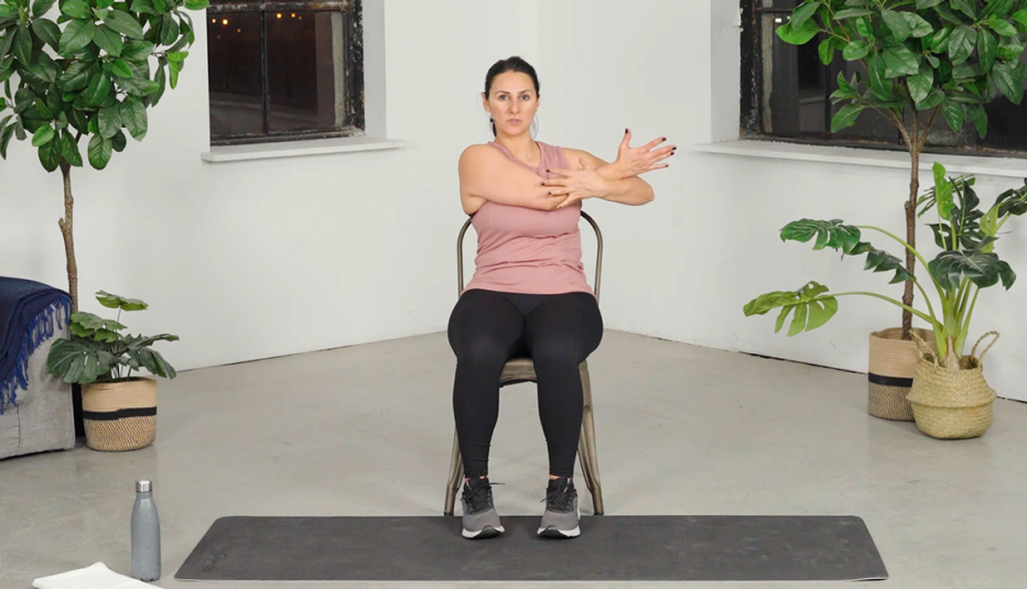 Shahnaz Mumeni sitting in chair on mat, stretching right arm across body with left arm pulling it