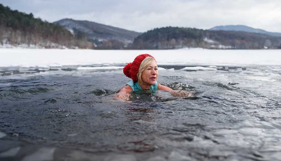 Cold water swimming and ice baths - are they good for you? woman wearing a winter hat swimming outdoors in an icy lake or river in winter with snow surrounding the water she is submerged up to her neck