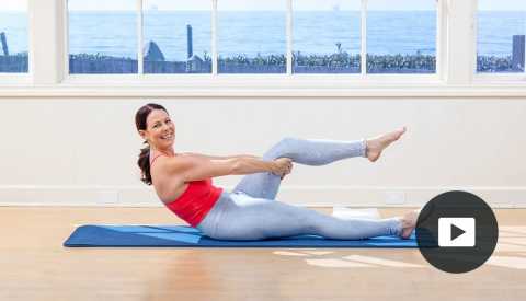 Pilates instructor Erin Wilson smiling in a Pilates pose on a mat in a studio with a window looking out onto the water, and video icon overlay