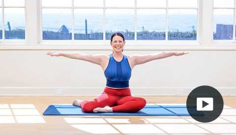 Pilates instructor Erin Wilson smiling in a Pilates pose on a mat in a studio with a window looking out onto the water, with video icon overlay