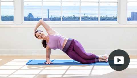 Pilates instructor Erin Wilson in a modified plank pose on a mat in a studio with a window looking out onto the water. video player button in right corner