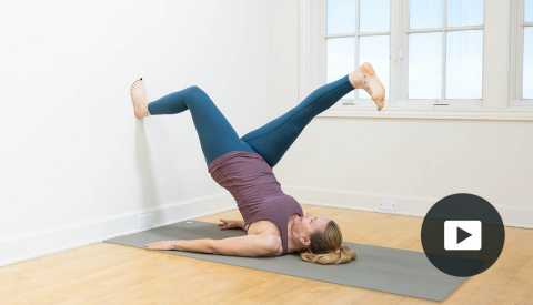 Pilates instructor Amy Havens lies on her back with one foot raised on a wall and the other stretched above her head. She is on a yoga mat. I window is in the background. Video player icon in lower right corner.