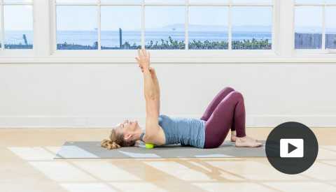 Pilates instructor Amy Havens lies on a yoga mat to perform a pose. In the background, windows show a water scene. Video player icon in the lower right corner.